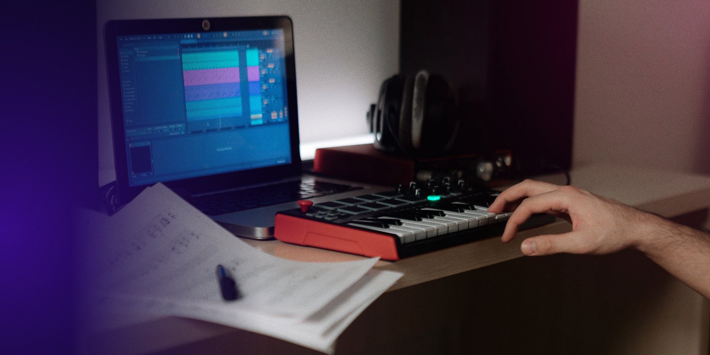 Dans un home studio, la main d'un artiste sur un petit clavier MIDI, l'écran de son logiciel et ses notes à côté.